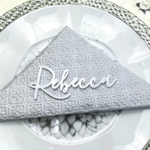 May include: A white, cursive name card with the name "Rebecca" on a gray, patterned napkin. The napkin is folded into a triangle shape and placed on a white plate with a silver rim.