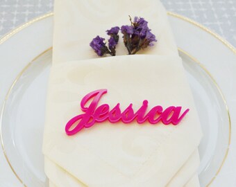 Peut inclure: Un marque-place en acrylique rose vif avec le prénom "Jessica" en écriture cursive, posé sur une serviette crème pliée. La serviette est sur une assiette blanche avec un bord doré, et une petite branche de fleurs violettes est insérée.