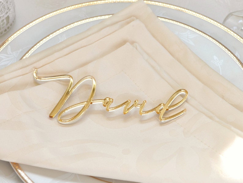 May include: Gold acrylic place card with the name "David" written in a cursive font.