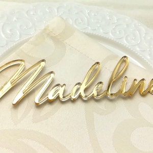 Peut inclure: Un marque-place doré en acrylique avec le prénom "Madeline" écrit en cursive. Il est posé sur une serviette crème, avec une assiette blanche décorative en arrière-plan. Idéal pour les mariages et événements.