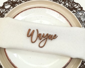 May include: A wooden place card with the name "Wayne" written in cursive script. The place card is sitting on a white napkin on a white plate.