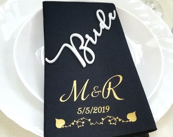 May include: Black napkin with gold lettering that says "M&R 5/5/2019" with a vine design. A white plastic sign that says "Bride" is on top of the napkin.
