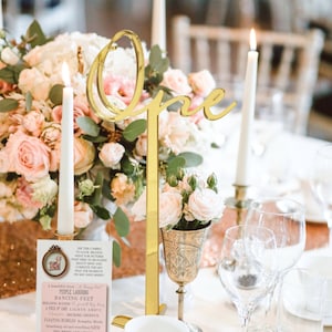 Pode incluir: Um número de mesa dourado com a palavra "One" em escrita cursiva, numa mesa com arranjos florais, velas e copos. Há também um pequeno cartão rosa com texto.