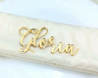 Puede incluir: Una placa con el nombre "Gloria" en acrílico dorado y fuente cursiva. La placa está colocada sobre una tela plegada de color blanco roto con un patrón sutil. Probablemente se utiliza para la decoración de mesas.