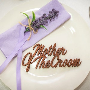May include: A white plate with a purple napkin tied with twine and a sprig of lavender. The plate also has a wooden sign that says "Mother of the Groom".