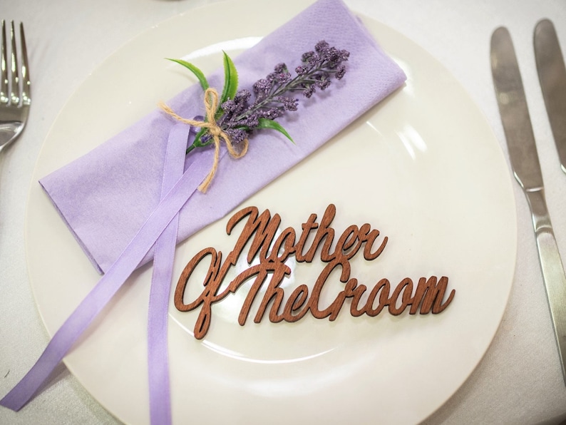 May include: A white plate with a purple napkin tied with twine and a sprig of lavender. The plate also has a wooden sign that says "Mother of the Groom".