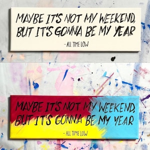 May include: A white canvas with black text that reads "MAYBE IT'S NOT MY WEEKEND, BUT IT'S GONNA BE MY YEAR - ALL TIME LOW". Below the canvas is a second canvas with the same text in black, but the background is painted in red, yellow, and blue.