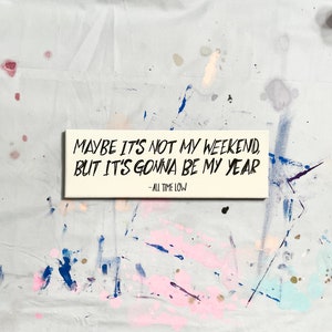 May include: A white wooden sign with black lettering that reads "Maybe it's not my weekend, but it's gonna be my year - All Time Low".