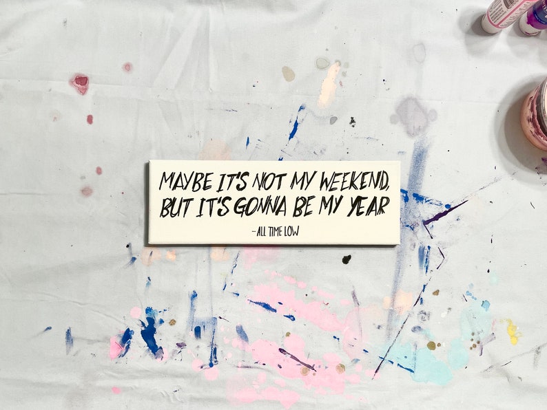May include: A white wooden sign with black lettering that reads "Maybe it's not my weekend, but it's gonna be my year - All Time Low".