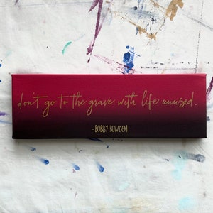 May include: A red and black canvas with the quote "don't go to the grave with life unused." in gold lettering. The quote is attributed to Bobby Bowden.