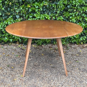 May include: A light brown, oval wooden table with three angled legs. The table has a natural wood grain finish. The table is set against a backdrop of green foliage and a gravel surface.