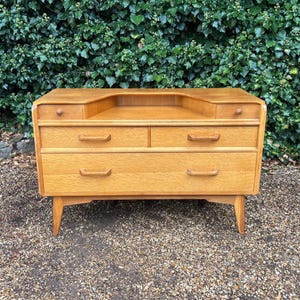 May include: A light brown wooden chest of drawers featuring a recessed top shelf, two small upper drawers with round knobs, two medium-sized drawers with horizontal handles, and one large bottom drawer with two horizontal handles, supported by four tapered legs.