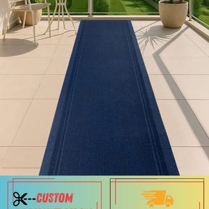 May include: A navy blue indoor/outdoor rug runner with a ribbed texture. The rug is rectangular and has two subtle stripes running down the length. The rug is on a tiled surface, with a chair and table in the background.