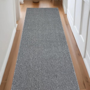 May include: A rectangular gray hallway runner rug with a textured surface. The rug is placed on a wooden floor, and the edges are finished with a thin, light-colored border. The runner extends down a hallway.