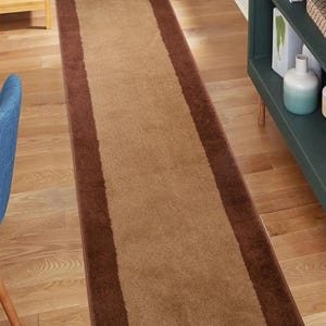 May include: A brown and beige runner rug with a brown border. The rug is made of a soft, plush material and has a subtle texture.