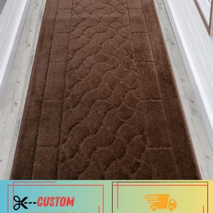 May include: A brown runner rug with a textured, patterned design. The rug features a central wavy pattern and a rectangular border. The rug is placed in a hallway with white trim and light-colored flooring. The rug is a custom size.