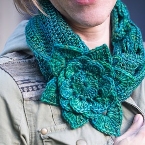 Peut inclure: Une écharpe en crochet bleu sarcelle avec un grand motif de fleur. L'écharpe est portée autour du cou d'une personne portant une veste verte.
