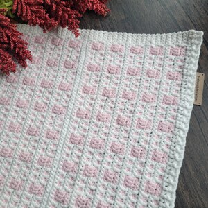 May include: A white and pink crocheted blanket with a square pattern. The blanket is made with a soft yarn and has a delicate texture.