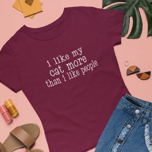May include: A maroon t-shirt with the slogan "i like my cat more than i like people" in white. The image also shows a denim skirt, sandals, and nail polish, all arranged on a pink surface.