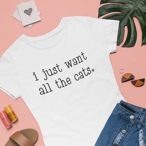 May include: White t-shirt with the text "i just want all the cats" in black. The shirt is paired with a denim skirt, brown sandals, and nail polish on a pink background. Decorative heart-shaped cards and earrings are also present.