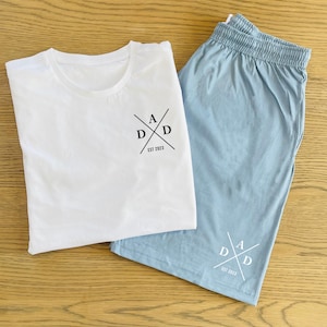 May include: A white t-shirt with a black logo that says "ADD EST 2023" and a pair of light blue shorts with a white logo that says "ADD EST 2023".