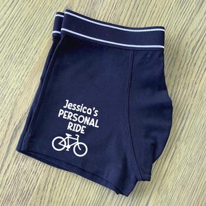 May include: Black boxer shorts with a white elastic waistband and a white graphic of a bicycle with the text "Jessica's Personal Ride".