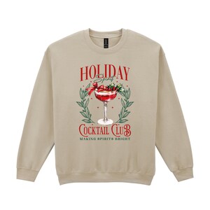 May include: A tan crewneck sweatshirt with a festive graphic. The graphic features a cocktail in a stemmed glass, surrounded by greenery and the words "Holiday Spirit Cocktail Club, Making Spirits Bright" in red and white.