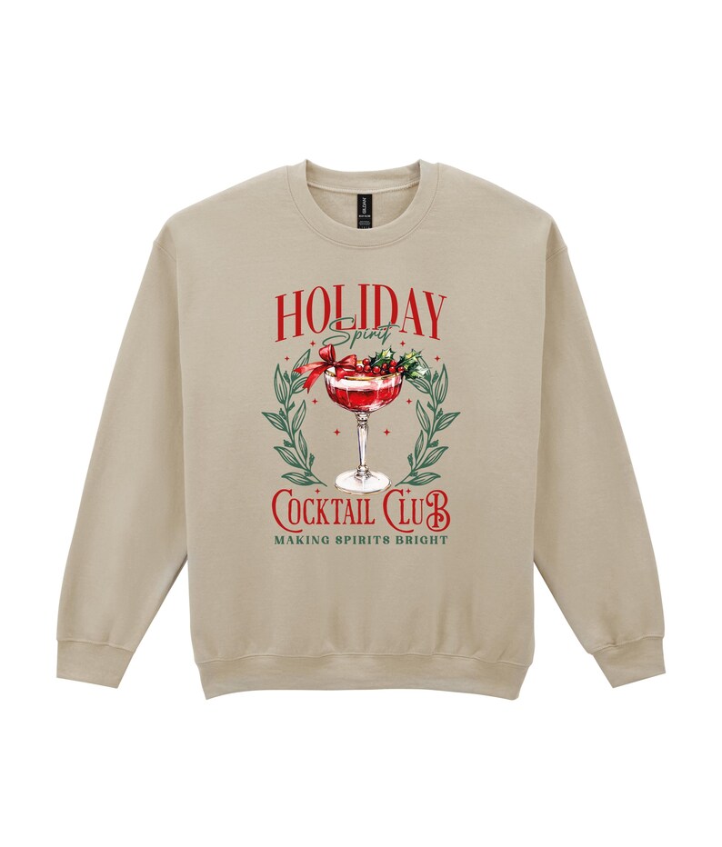 May include: A tan crewneck sweatshirt with a festive graphic. The graphic features a cocktail in a stemmed glass, surrounded by greenery and the words "Holiday Spirit Cocktail Club, Making Spirits Bright" in red and white.