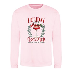 May include: A light pink sweatshirt with a graphic that reads "Holiday Spirit Cocktail Club" in red and white. The graphic features a cocktail in a stemmed glass, surrounded by a wreath of green leaves and berries.