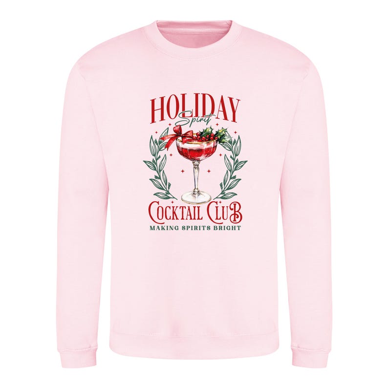 May include: A light pink sweatshirt with a graphic that reads "Holiday Spirit Cocktail Club" in red and white. The graphic features a cocktail in a stemmed glass, surrounded by a wreath of green leaves and berries.