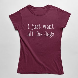 May include: Burgundy t-shirt with the text "i just want all the dogs" in white. The word "dogs" has a paw print graphic. The shirt is a crew neck and has short sleeves.