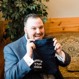 May include: A man in a blue suit is holding up a pair of dark blue underwear with the text "PROPERTY OF Mrs. Brake  SEPTEMBER 2018" embroidered on them.