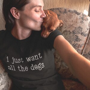 May include: Black t-shirt with the text "i just want all the dogs" in white. A small brown dog rests on the shoulder of a person wearing the shirt. The person is kissing the dog. The shirt is made of a soft material.