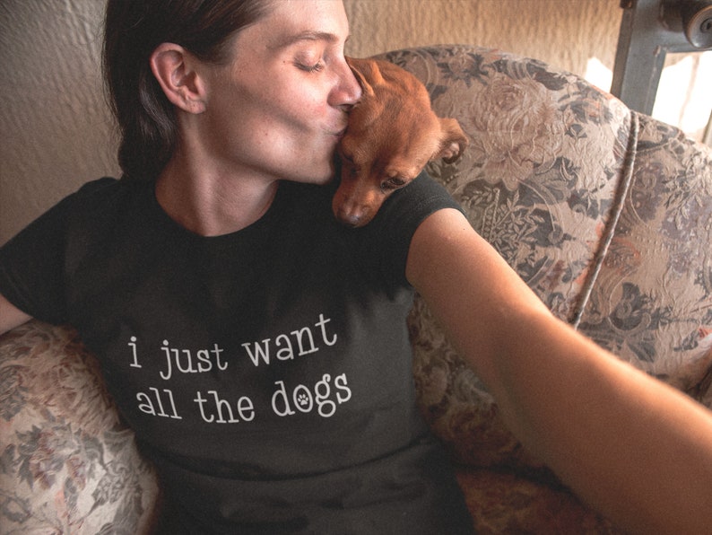 May include: Black t-shirt with the text "i just want all the dogs" in white. A small brown dog rests on the shoulder of a person wearing the shirt. The person is kissing the dog. The shirt is made of a soft material.