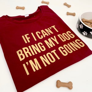 May include: A burgundy t-shirt with the gold text: "IF I CAN'T BRING MY DOG I'M NOT GOING." A dog bowl with paw prints contains dog biscuits. Several dog treat biscuits are scattered around the shirt and bowl.