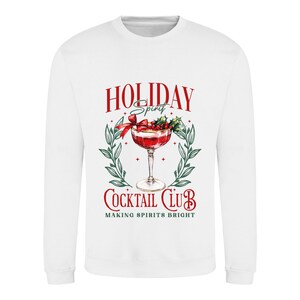May include: White sweatshirt with a festive cocktail graphic. The design features a red cocktail with a bow, holly, and the words "HOLIDAY Spirit" and "COCKTAIL CLUB" in red, with green accents. The text "MAKING SPIRITS BRIGHT" is also included.