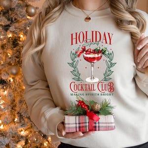 May include: A beige sweatshirt with a festive graphic that reads "HOLIDAY Spirit COCKTAIL CLUB" with a cocktail illustration. The sweatshirt is paired with a plaid gift box with a red bow and greenery.