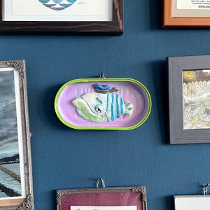 Könnte beinhalten: Ein skurriles Wandkunstwerk mit einer Fischskulptur in einer wiederverwendeten Konservendose. Der Fisch ist weiß mit blauen Streifen und einem blauen Hut, vor einem rosa Hintergrund mit einem grünen Rand. Das Kunstwerk ist an einer blauen Wand befestigt.