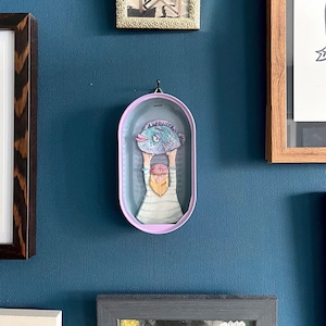 Könnte beinhalten: Ein skurriles Wandbild, das eine Figur zeigt, die einen großen blauen Fisch hochhält. Das Kunstwerk befindet sich in einem hellvioletten ovalen Rahmen mit blauem Innenraum. Die Figur hat einen langen weißen Bart und trägt ein gestreiftes Hemd.