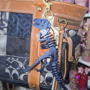 May include: A dark blue dinosaur skeleton keychain hanging from a brown and blue patchwork handbag. The handbag has gold hardware and brown leather trim. The keychain is attached with a gold clasp.