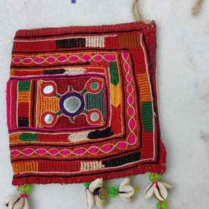 Può includere: Una piccola borsa quadrata rossa con ricami colorati e specchi. La borsa ha un anello di spago in cima ed è decorata con conchiglie bianche e perline verdi.