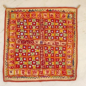May include: A square, embroidered textile with a vibrant, geometric design. The central pattern features red squares with yellow, green, and blue accents. The border is a contrasting orange and red. The textile has a beige backing and a loop at each corner.