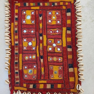 May include: A red, yellow and black embroidered textile with a geometric pattern and small mirrors. The textile is bordered with white seashells.