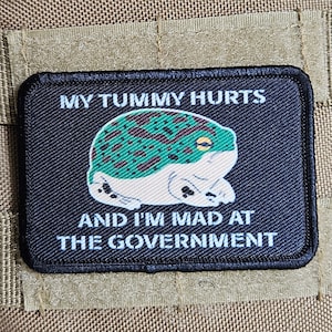 Peut inclure: Un sac à dos tactique beige avec deux patchs. Le patch du haut est un drapeau américain noir et blanc. Le patch du bas est noir avec une grenouille verte et le texte "MY TUMMY HURTS AND I'M MAD AT THE GOVERNMENT".