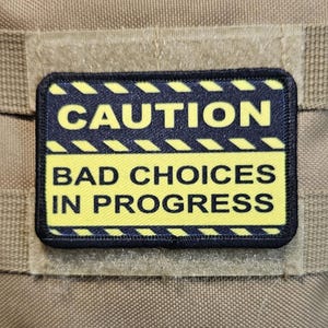 Op de afbeelding: Een bruine tactische vest met een zwart-witte Amerikaanse vlag patch en een gele en zwarte waarschuwingspatch met de tekst "CAUTION BAD CHOICES IN PROGRESS".