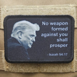 以下が含まれることがあります： 男性の顔の白い画像と「No weapon formed against you shall prosper - Isaiah 54:17」というテキストが書かれた黒いパッチ。