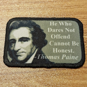 May include: A black and white embroidered patch with a portrait of Thomas Paine and the quote "He Who Dares Not Offend Cannot Be Honest. -Thomas Paine"