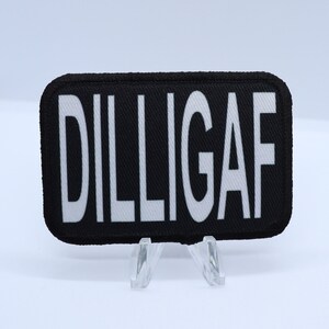 May include: Black rectangular patch with the white text "DILLIGAF". The patch has rounded corners and is displayed on a clear stand against a white background. The text is in a bold, sans-serif font.