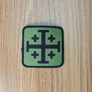 Könnte beinhalten: Ein grüner quadratischer Patch mit einem schwarzen Kreuz mit vier kleineren Kreuzen an jedem Arm. Der Patch ist aus Stoff und hat einen schwarzen Rand.