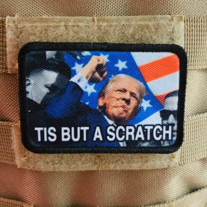 May include: A rectangular patch with the text "TIS BUT A SCRATCH" and an image of a person in front of an American flag. The patch is attached to a tan tactical vest.
